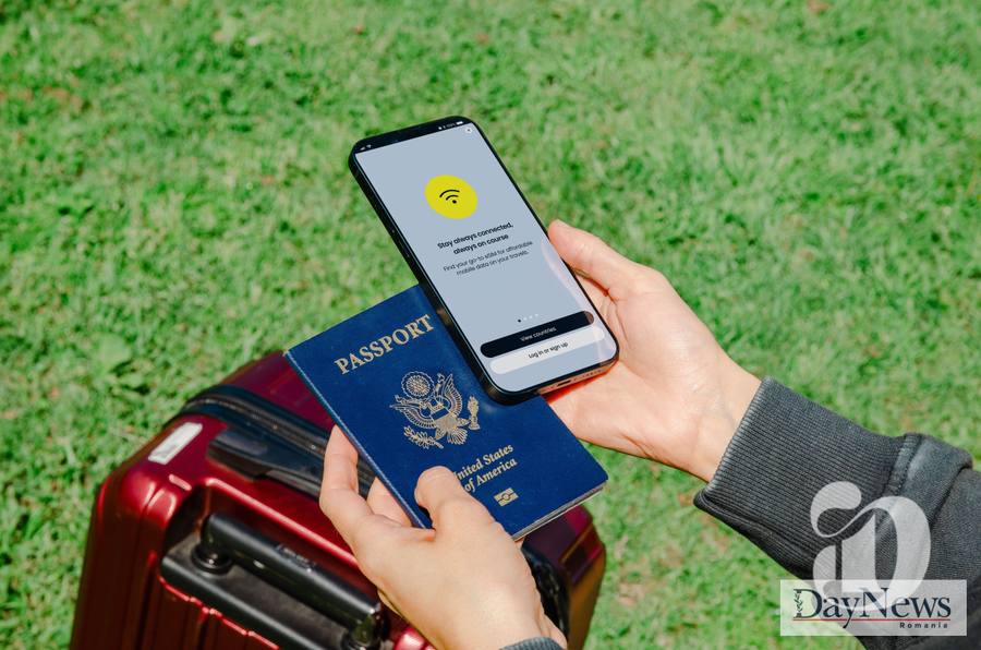 O persoană ține un pașaport american și un smartphone deschis pe iarbă verde, lângă un troler roșu