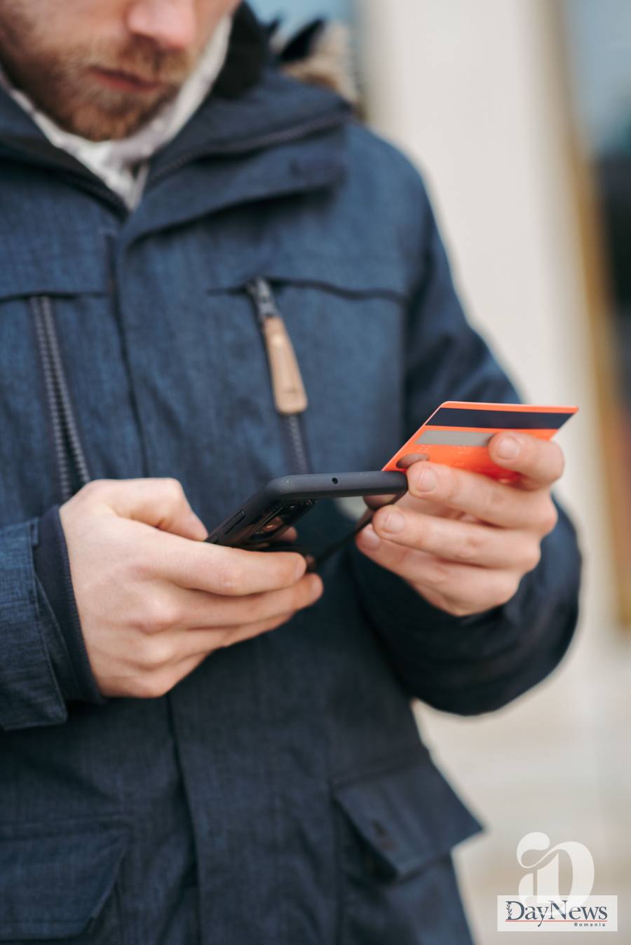 Un bărbat cu geacă de iarnă albastră ține un telefon și un card portocaliu în mâini